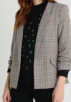 Femme portant un blazer marron et blanc à carreaux par-dessus un pull noir avec de petits motifs floraux verts et un pantalon noir.