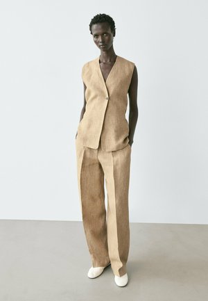 Modella che indossa un gilet beige in lino senza maniche con un bottone e pantaloni larghi abbinati, in piedi con le mani nelle tasche davanti a una parete bianca.