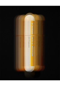 Tube de mascara Maybelline en blanc et jaune, forme cylindrique, avec une prise texturée et un logo réfléchissant sur un fond sombre.
