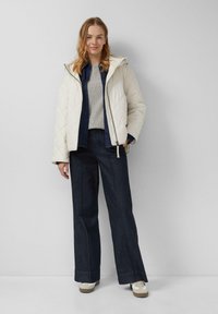 Vrouw die staat met een crèmekleurige gewatteerde jas over een grijze sweater en donkerblauwe wijde broek met witte sneakers tegen een effen muur.