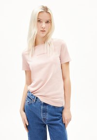 Lichtroze katoenen T-shirt met korte mouwen, ronde halslijn en voorop de tekst "ARMEDANGELS." Gecombineerd met blauwe denim jeans.