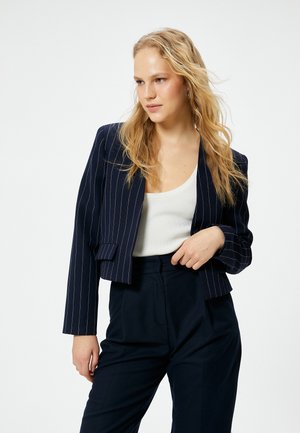 Marineblauwe pinstripe blazer met een cropped ontwerp, nauwsluitende pasvorm en gestructureerde schouders. Gerdraagd over een wit tanktop, aangevuld met donkere broek.