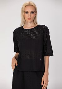 Haut noir avec un design en œillets, manches courtes, encolure ronde, tissu léger, présentant un motif texturé et une coupe décontractée.