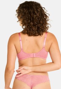 Soutien-gorge en dentelle rose avec des bretelles réglables et une fermeture arrière à crochets. Des motifs de feuilles complexes créent un design texturé.