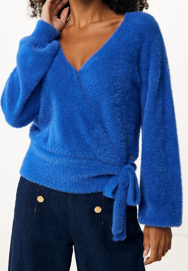Femme portant un pull enveloppant pelucheux bleu vif à manches longues noué à la taille, assorti à un pantalon bleu foncé avec des boutons dorés.