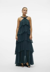 Tirkizna slojevita chiffon haljina s halter izrezom, koja ima naborane slojeve, lepršavi dizajn i cvjetni naglasak oko vrata.