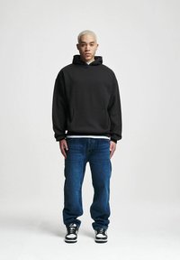 Felpa nera con cappuccio e tasca frontale, indossata sopra una maglietta bianca; abbinata a jeans blu e sneakers bianco e nere.