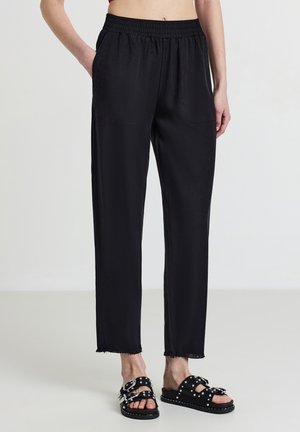Person in schwarzen elastischen Hosen mit fransigen Säumen und schwarzen besetzten Slide-Sandalen vor einem schlichten Hintergrund.