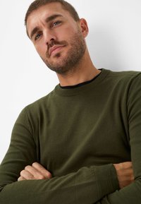Maglione a coste di colore verde oliva con scollo rotondo, polsini a coste e texture liscia. Il modello ha le braccia incrociate, evidenziando il design del maglione.