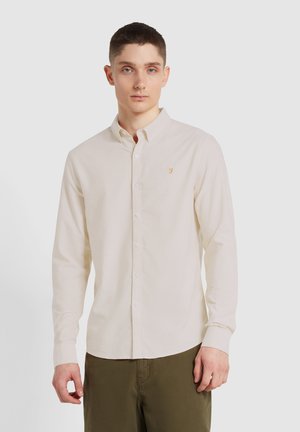 Chemise à manches longues de couleur beige clair avec un col à boutons, dotée de boutons blancs et d'un petit logo orange sur la poitrine gauche. Texture lisse.