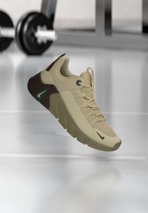 Béžovo-hnedá športová obuv s hrubou podrážkou, logo Nike na boku a na špičke, zavesená nad podlahou posilňovne s posilňovacím vybavením v pozadí.