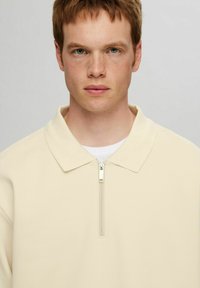 Beige polo tričko na zip s texturovanou tkaninou, které má klasický límeček a stříbrný zip na krku.