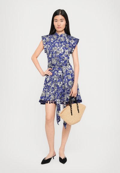 Floral print dress in deep blue with short ruffled sleeves, belted waist, and tassel hem. Model holds a woven straw bag with black handles.