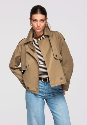 Kvinde med glat hårover trukket tilbage, iført gyldne hoop-øreringe, en beige cropped trenchcoat over en sort-hvid stribet skjorte og lyseblå jeans.