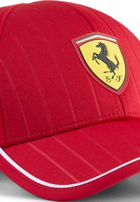 Czerwona czapka z pionowymi żeberkami, z żółtą naszywką z logo Ferrari i białym akcentem wzdłuż ronda.