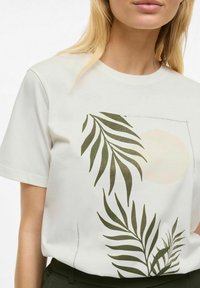 Valkoinen lyhythihainen t-paita, jossa on vihreitä lehtipainatuksia ja vaalean kehämäinen graafinen kuva, valmistettu pehmeästä materiaalista ja rentoa istuvuutta.