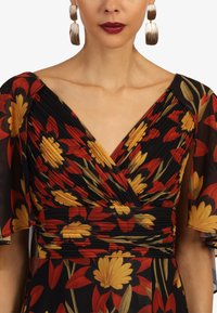 Schwarzes Blumenkleid mit roten und gelben Blumenmustern, gefälteltem Oberteil und kurzen, flatternden Ärmeln, kombiniert mit silbernen Statement-Ohrringen.