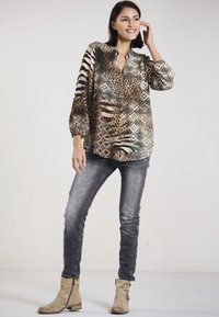 Woman smiling with short dark hair, wearing a patterned animal print blouse, grey jeans, and beige ankle boots, standing against a plain background.