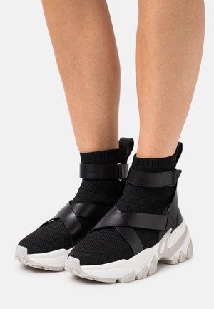Jambes portant des baskets style chaussettes noires avec des semelles blanches épaisses, des lanières en cuir noir croisant le pied et entourant la cheville.