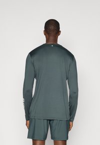 Dlhý rukáv, tmavozelené športové tričko vyrobené z hladkého materiálu, s okrúhlym výstrihom a nenápadnou detailnou logom na zadnej strane.