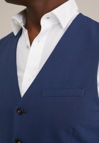 Námořnický modrý vest s hladkou texturou, vybavený klopou, dvěma knoflíky a jednou náprsní kapsou, nošený přes bílou košili s límečkem.