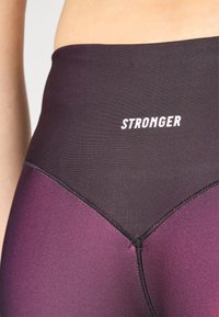 Svarte og mørklilla høytekkede leggings med en jevn tekstur, med en hvit "STRONGER" logo og kontrasterende sømdetaljer.