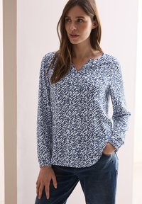 Blaue und weiße gemusterte Bluse mit V-Ausschnitt, langen Ärmeln und lockerer Passform. Der Stoff ist leicht und hat eine glatte Textur.