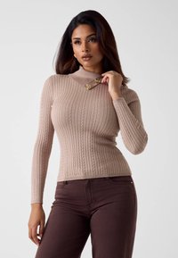 Maglione beige a costine con collo alto e bottoni decorativi dorati sul colletto. Abbinato a pantaloni aderenti marrone scuro.