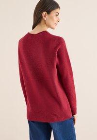 Maglione rosso lavorato a maglia con colletto rotondo, polsini e orlo a costine. Texture morbida, taglio leggermente oversize, indossato sopra jeans blu scuro.