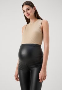 Beige ribbad ärmlös topp i kombination med svarta leggings i fusk läder för gravida, med en slät textur och en figurnära form runt magen.