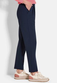 Persoon in marineblauwe tapschrift broek met handen in de zakken en beige sneakers met roze en witte accenten op een lichte achtergrond.