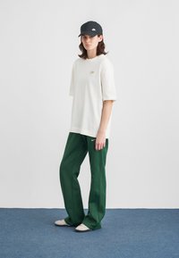 Άτομο που φοράει ένα μαύρο καπέλο Nike, λευκή oversized μπλούζα, σκούρα πράσινα παντελόνια με λογότυπο Nike και ανοιχτό γκρι παπούτσια σε μπλε χαλί με φόντο λευκό τοίχο.