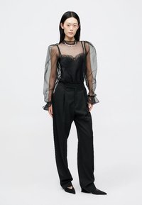 Μαύρη διαφανής μπλούζα με φουσκωτά μανίκια και δαντέλα, συνδυασμένη με ψηλόμεσες μαύρες παντελόνες και μυτερά μαύρα παπούτσια. Απλή κοσμήματα ως αξεσουάρ.