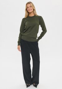 Jersey de color verde oliva con puños y dobladillo acanalados, combinado con pantalones sueltos negros y tacones negros de punta.