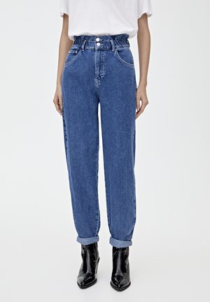 Jeans de mezclilla azul de tiro alto con un ajuste relajado, que cuentan con una cinturilla elástica fruncida y puños enrollados, combinados con botas negras hasta el tobillo.