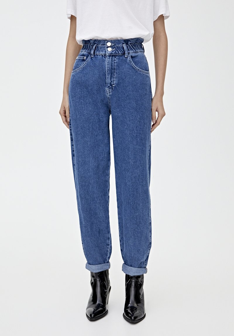 Jeans de mezclilla azul de tiro alto con un ajuste relajado, que cuentan con una cinturilla elástica fruncida y puños enrollados, combinados con botas negras hasta el tobillo.
