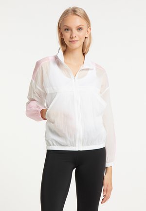 Jonge vrouw die een witte, lichtgewicht jas met ritssluiting draagt met roze accenten en zwarte legging, staand met één hand in haar zak.
