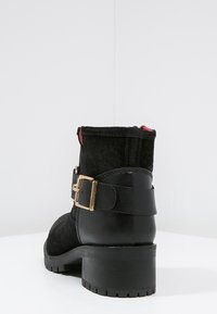 Black ankle boot made of suede and leather, featuring a gold hardware buckle and a textured rubber sole. Short, stacked heel with a smooth finish.