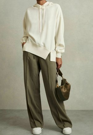 Personne portant un sweat à capuche crème oversize, un pantalon large olive, des baskets blanches, tenant un sac à main olive par sa anse.