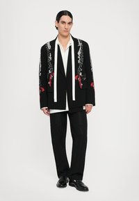 Schwarze Jacke mit weißen Revers und floralem Stickerei, die rote Akzente aufweist, kombiniert mit einem weißen Hemd und schwarzen strukturierten Hosen.