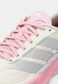 Ελαφρύ αθλητικό παπούτσι με επάνω μέρος από δίχτυ σε εκρού και ροζ, με ροζ κορδόνια, τρεις γκρι ρίγες και μια ελαφρώς μαξιλαρωμένη σόλα.