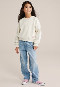 Felpa di colore crema, con texture e scollatura a V, abbinata a jeans in denim chiaro, morbidi e larghi, e scarpe da ginnastica rosa. Piccolo logo stampato sulla felpa.