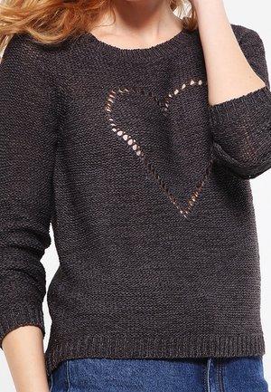Pull noir en maille tricotée avec un motif en maille ajourée en forme de cœur sur la poitrine, porté avec un jean bleu.
