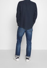 Mørkeblå langærmet skjorte med rundet kant, parret med lyseblå jeans. Hvide sneakers med sorte detaljer fuldender outfittet.
