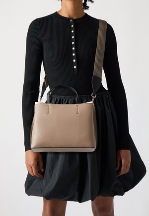 Sac à main en cuir beige avec un design structuré, de courtes anses noires et une bandoulière tissée amovible. Porté avec une robe noire à côtes.