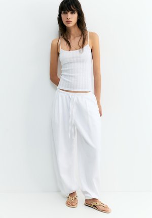 Femme aux cheveux foncés portant un haut blanc sans manches côtelé, un pantalon blanc ample à cordon et des sandales beiges, debout contre un mur blanc.