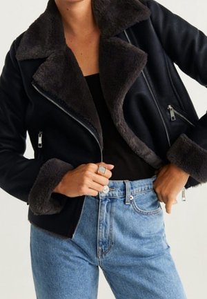 Person iført en sort shearling-foret jakke med lynlåse og lyseblå højttaljede jeans, der viser hænder, der justerer jakken og jeansene.