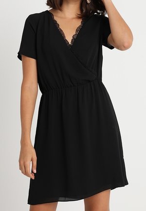 Woman wearing a short black wrap dress with lace trim on the V-neckline and short sleeves.