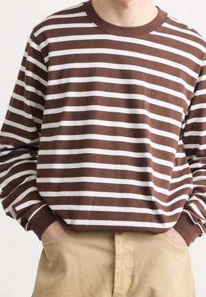 Osoba ubrana w brązowo-biały długorękawowy T-shirt w poziome paski z inicjałami "MN" na piersi, w połączeniu z beżowymi spodniami, ręce w kieszeniach.