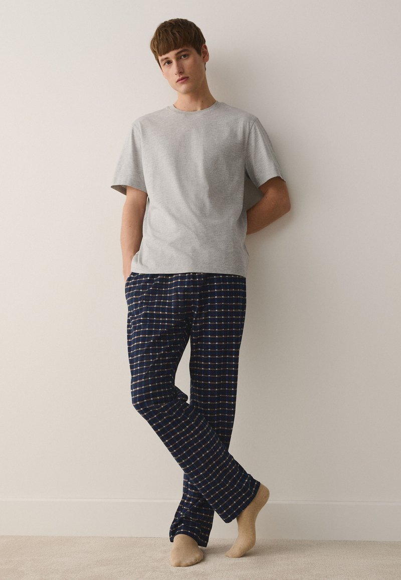 Jeune homme aux cheveux bruns portant un t-shirt gris à manches courtes et un pantalon de pyjama bleu marine à carreaux, debout pieds nus contre un mur blanc.
